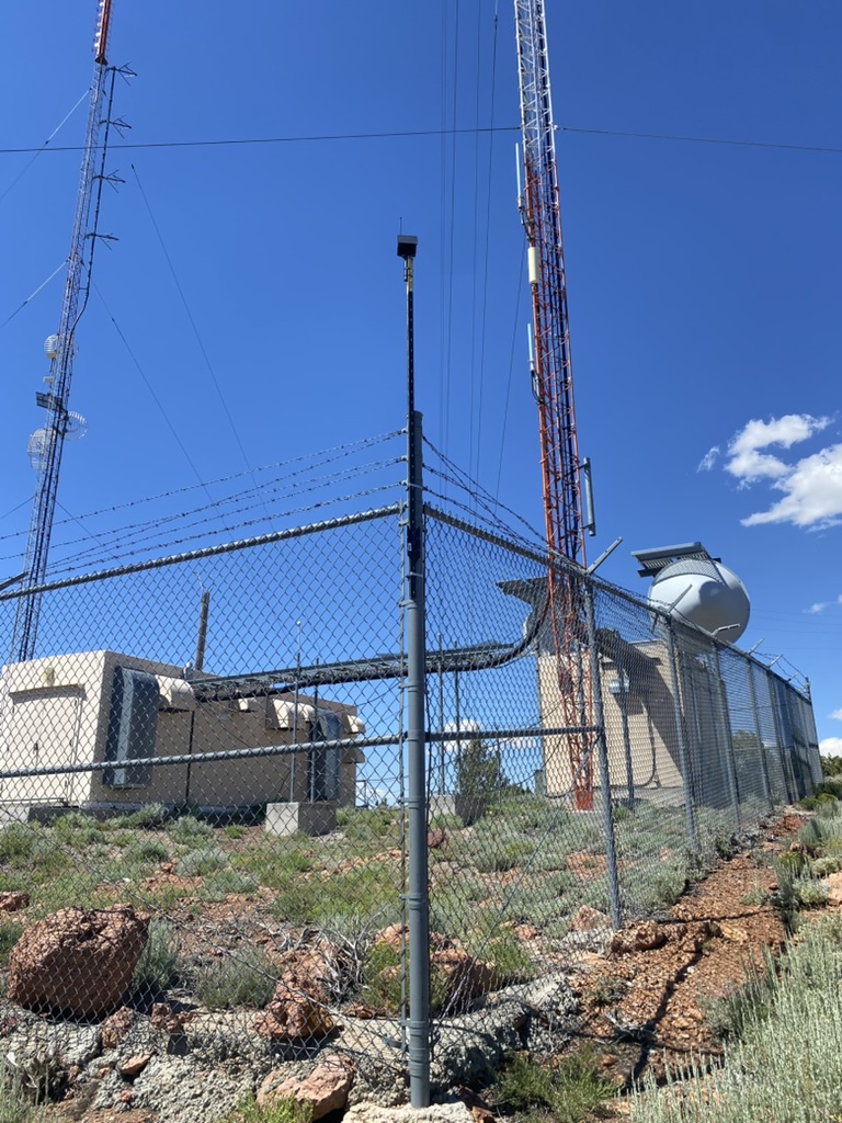 MeshCore node on a T-stake mast hose-clamped to a fence pole at the McClellan Peak compound